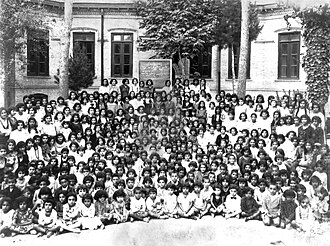 Fotografía en blanco y negro de varias docenas de niñas sentadas frente a un edificio escolar