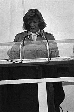 alt=A black and white photo of a woman in her fifties making a speech behind a podium and microphone